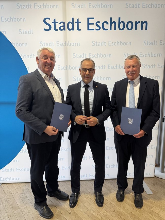 Karlheinz Gritsch, Bürgermeister Adnan Shaikh Helmut Bauch vor einer Stadt-Eschborn-Fotowand. Die Stadträte halten blaube Urkundenmappen mit weißem Stadtwappen in den Händen.