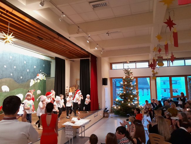 Auf der Bühne des Gemeindehauses tritt eine Gruppe Kinder in weißen T-Shirts und Nikolausmützen auf. Rechts schauen die Gäste der Feier gespannt zu. In der Mitte des Bildes steht ein erleuchteter Weihnachtsbaum.