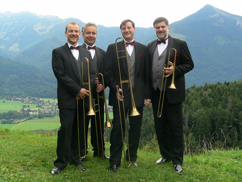 Das Posaunenquartett steht in Anzug und roter Fliege mit Instrumenten vor einer Berglandschaft.