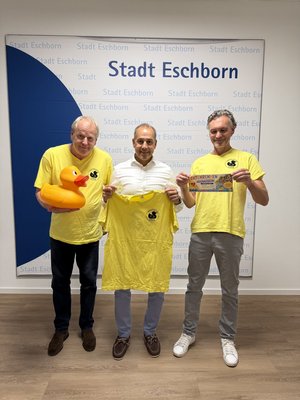Stephan Aders, Adnan Shaikh und Frank Dömski mit einer großen gelben Badeente und gelben T-Shirts vor der Fotowand mit großem, dunkelblauem Halbkreis links und dem Schriftzug Stadt Eschborn im Hintergrund