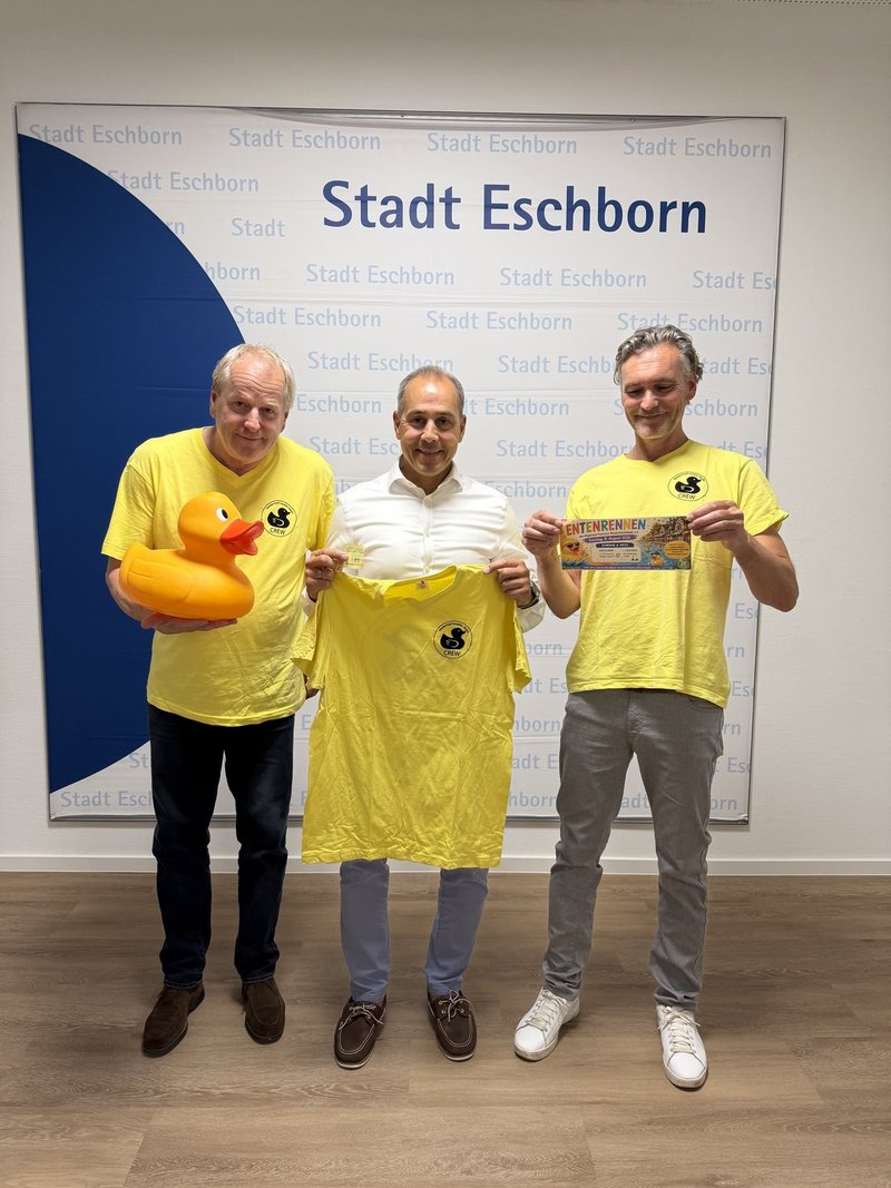 Stephan Aders, Adnan Shaikh und Frank Dömski mit einer großen gelben Badeente und gelben T-Shirts vor der Fotowand mit großem, dunkelblauem Halbkreis links und dem Schriftzug Stadt Eschborn im Hintergrund