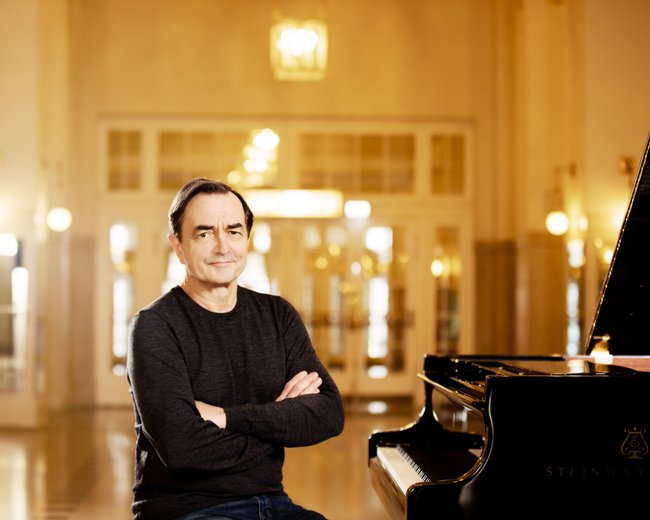 Pianist Pierre-Laurent Aimard sitzt mit verschränkten Armen und schwarz gekleidet neben einem schwarzen Klavier.