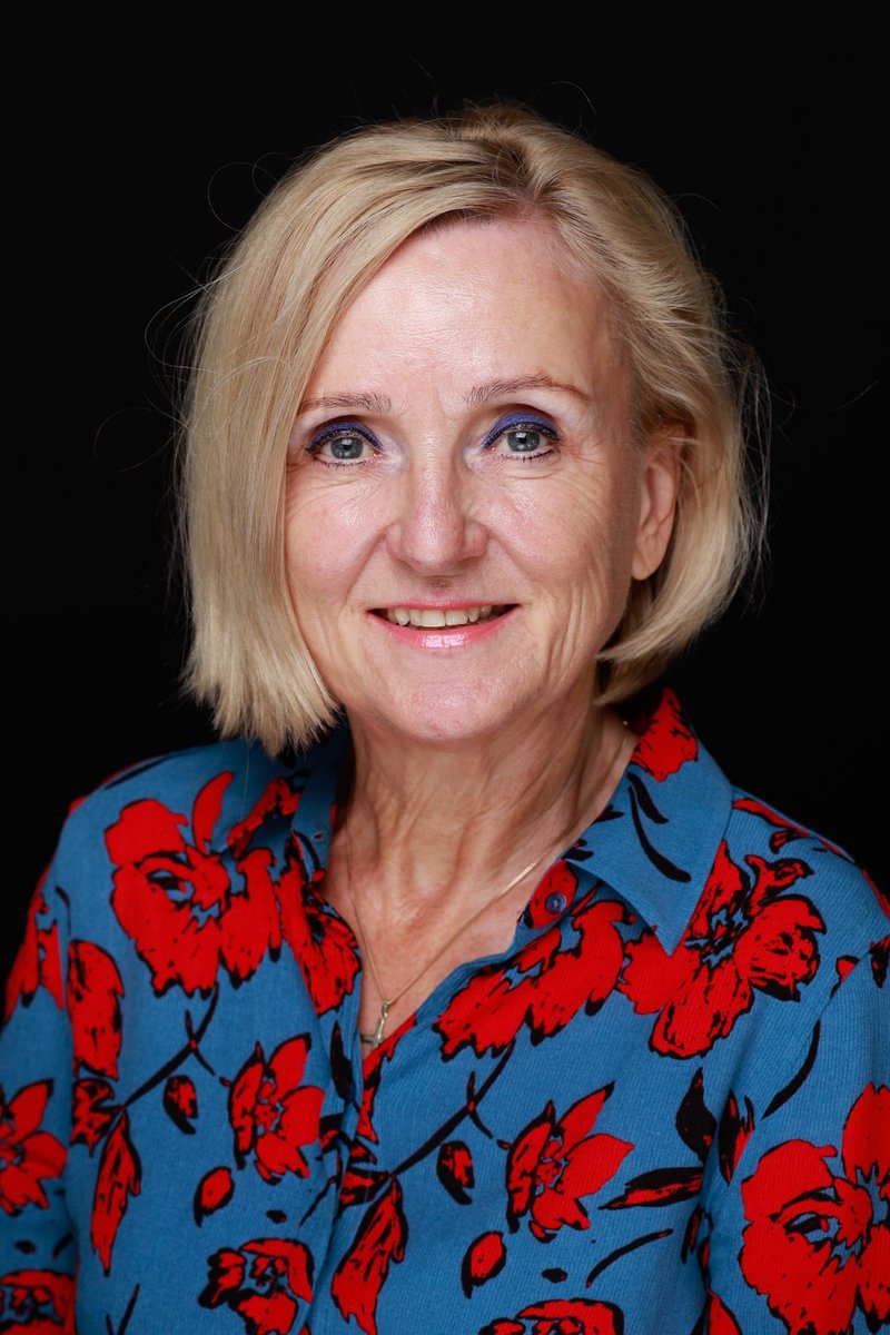 Eva Heymann hat blonde Haare. Sie trägt eine Bluse mit roten Blumen auf blauem Untergrund.