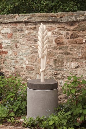 Skulptur einer hellen Ähre auf einem grauen Sockel vor einer Natursteinmauer