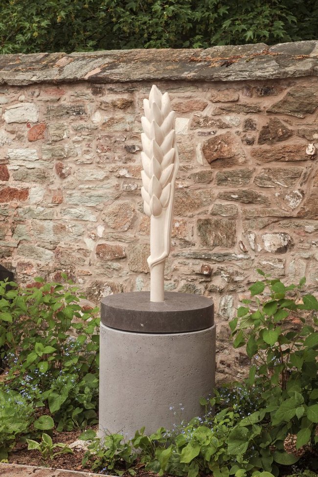 Skulptur einer hellen Ähre auf einem grauen Sockel vor einer Natursteinmauer