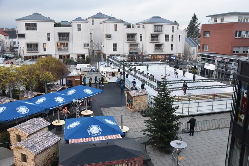 Blick von oben auf den Rathausplatz. Rechts sieht man die Eisbahn, links die hellbraunen Hütten unter blauen Schirmen.