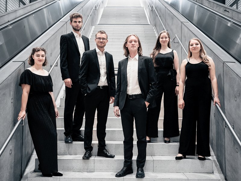 Drei Männer und drei Frauen in eleganter, dunkler Kleidung stehen auf einer hellen Treppe.