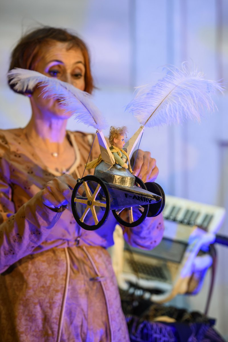 Eine Frau mit kurzen braunen Haaren hält einen kleinen Wagen mit Federn als Flügel in der Hand. Darin sitzt eine Figur des kleinen Prinzen.