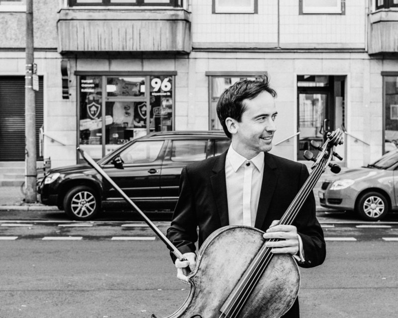 Schwarzweiß-Foto von Jean-Guihen Queyras mit Cello. Er steht auf einer Straße und blickt nach rechts.