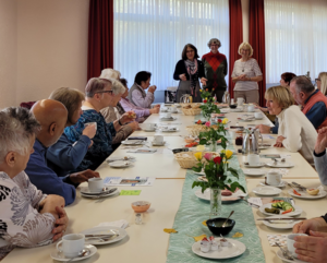 Seniorinnen und Senioren sitzen an einer hell gedeckten Frühstückstafel. Vor Kopf stehen Bärbel Grade, Esther Surkau und Christa Rochell.