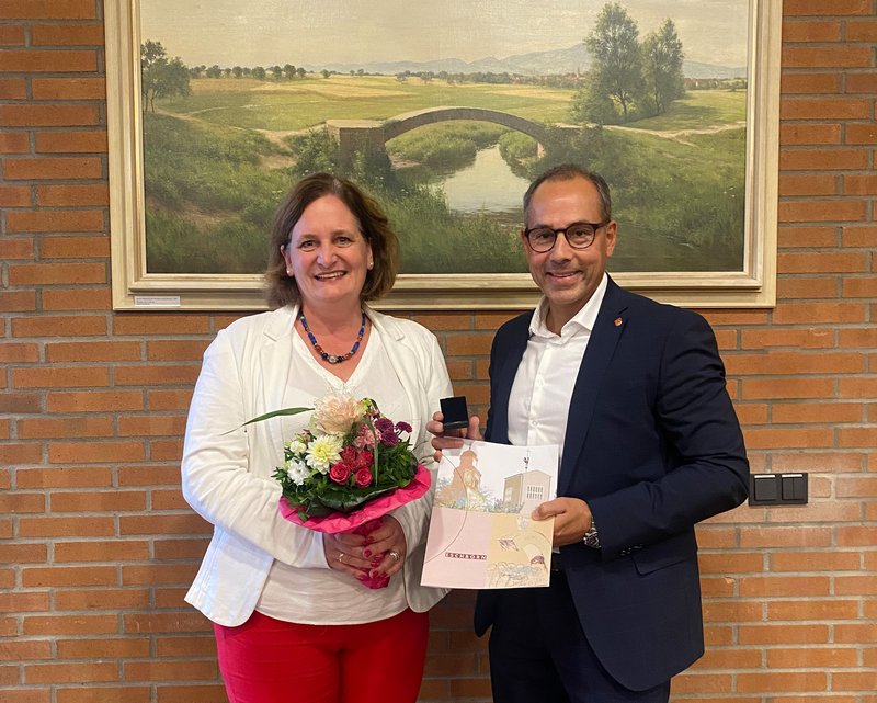 Frau Greiner und Bürgermeister Shaikh stehen vor einem Bild an einer Ziegelwand. Sie hält einen Blumenstrauß, er die Urkunde.