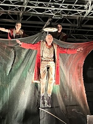 Till Eulenspiegel im roten Mantel mit ausgebreiteten Armen vor einem schwarzen Tuch. Es sieht aus als würde er schweben.