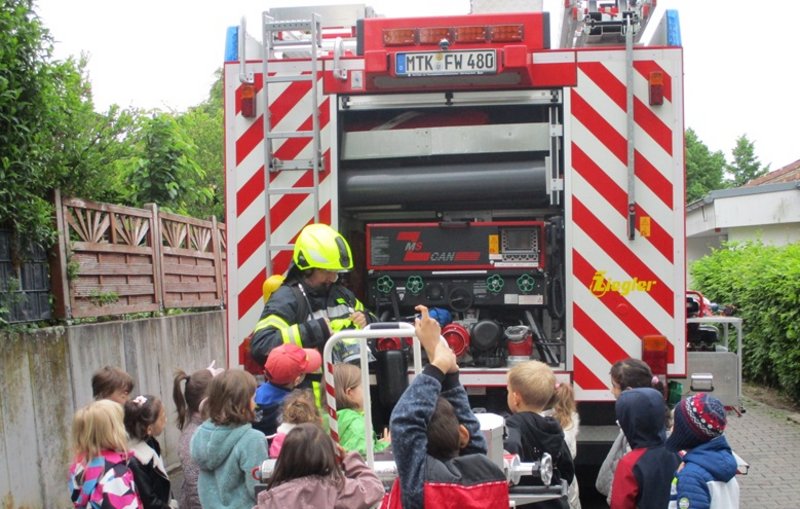 Eine Person in Schutzkleidung der Feuerwehr mit einer Kindergruppe vor einem Rettungswagen.