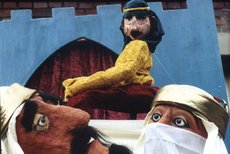Zwei Pappmaché-Figuren in weißer Kleidung blicken zu einer dritten hoch, die ein gelbes Oberteil und eine blaue Kufiya trägt.