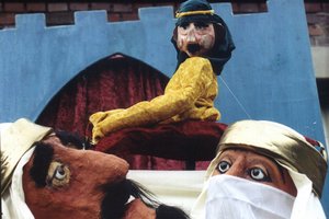 Zwei Pappmaché-Figuren in weißer Kleidung blicken zu einer dritten hoch, die ein gelbes Oberteil und eine blaue Kufiya trägt.