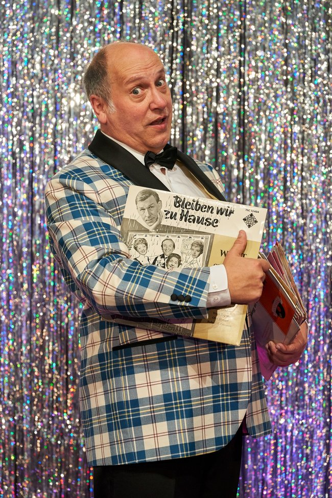 Willi Wunder im karierten Sakko vor silbernem Glitzervorhang. In der Hand hält er Platten.