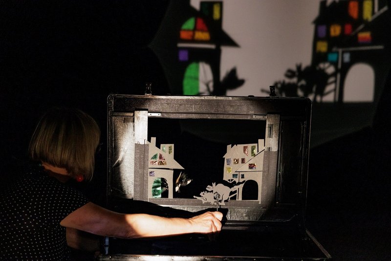 Ein heller Lichtkegel scheint auf eine Schattentheater-Szene. Rechts unten ist ein Unterarm einer Person zu sehen, die eine Figur hält.