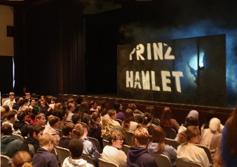 Jugendliche sitzen vor der Bühne, dort steht "Prinz Hamlet" in weißer Schrift auf schwarzem Grund.