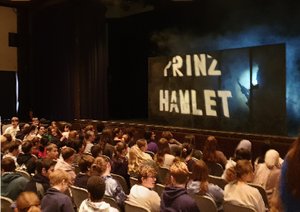 Jugendliche sitzen vor der Bühne, dort steht "Prinz Hamlet" in weißer Schrift auf schwarzem Grund.