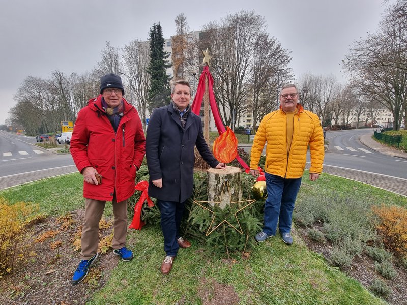 Links steht vor dem Adventskranz auf dem Kreisel Hans Peter Meyer in einer roten Jacke, rechts daneben Markus von Sternheim in einem dunkelblauen Mantel, er hat eine Hand auf der Holzflamme liegen. Rechts von ihm steht Udo Gauf in einer gelben Jacke.