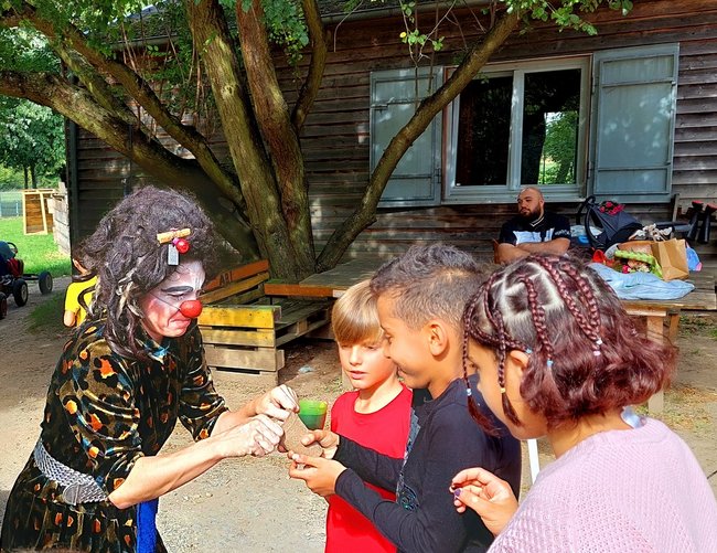 Eine Clownsfrau mit roter Plastiknase zeigt zwei Jungen und einem Mädchen einen Trick.