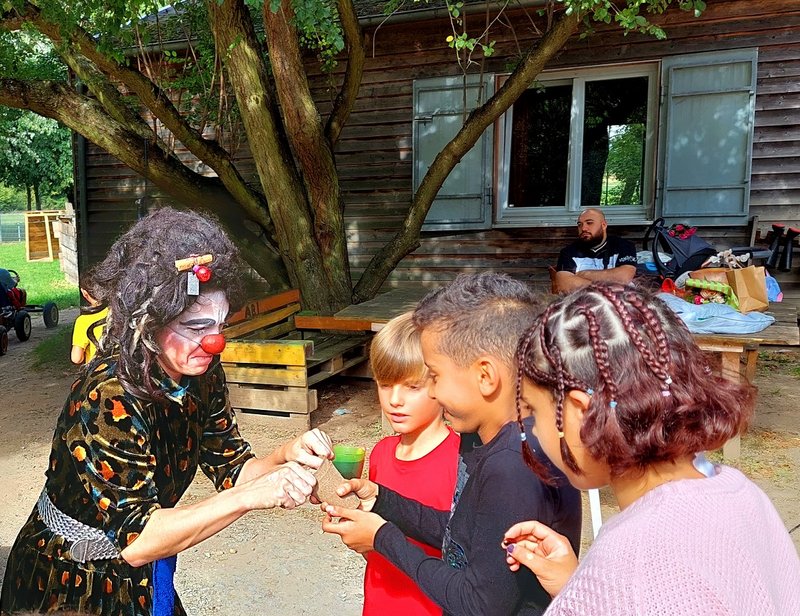 Eine Clownsfrau mit roter Plastiknase zeigt zwei Jungen und einem Mädchen einen Trick.