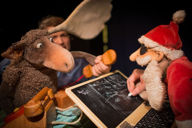 Puppentheater: links ein Elch mit braunem Fell, rechts schreibt ein Weihnachtsmann in weiß-roter Kleidung auf eine Tafel