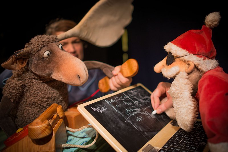 Puppentheater: links ein Elch mit braunem Fell, rechts schreibt ein Weihnachtsmann in weiß-roter Kleidung auf eine Tafel