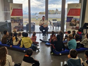 Stefan Leder sitzt auf einem Stuhl zwischen zwei Rollups mit Infos zur Feuerwehr. Er hält ein Buch in der Hand. Vor ihm sitzen Kinder auf blau-schwarzen Sitzkissen.