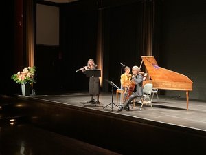 Auf der Bühne steht eine Flötistin, ein Cellist und eine Pianistin sind rechts und in der Mitte zu sehen