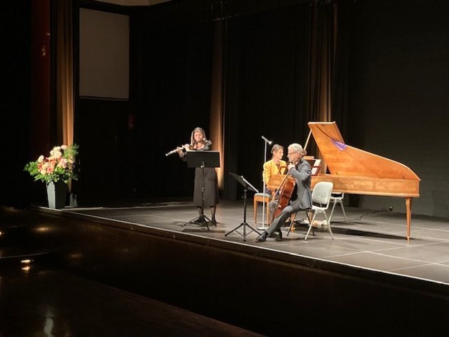 Auf der Bühne steht eine Flötistin, ein Cellist und eine Pianistin sind rechts und in der Mitte zu sehen