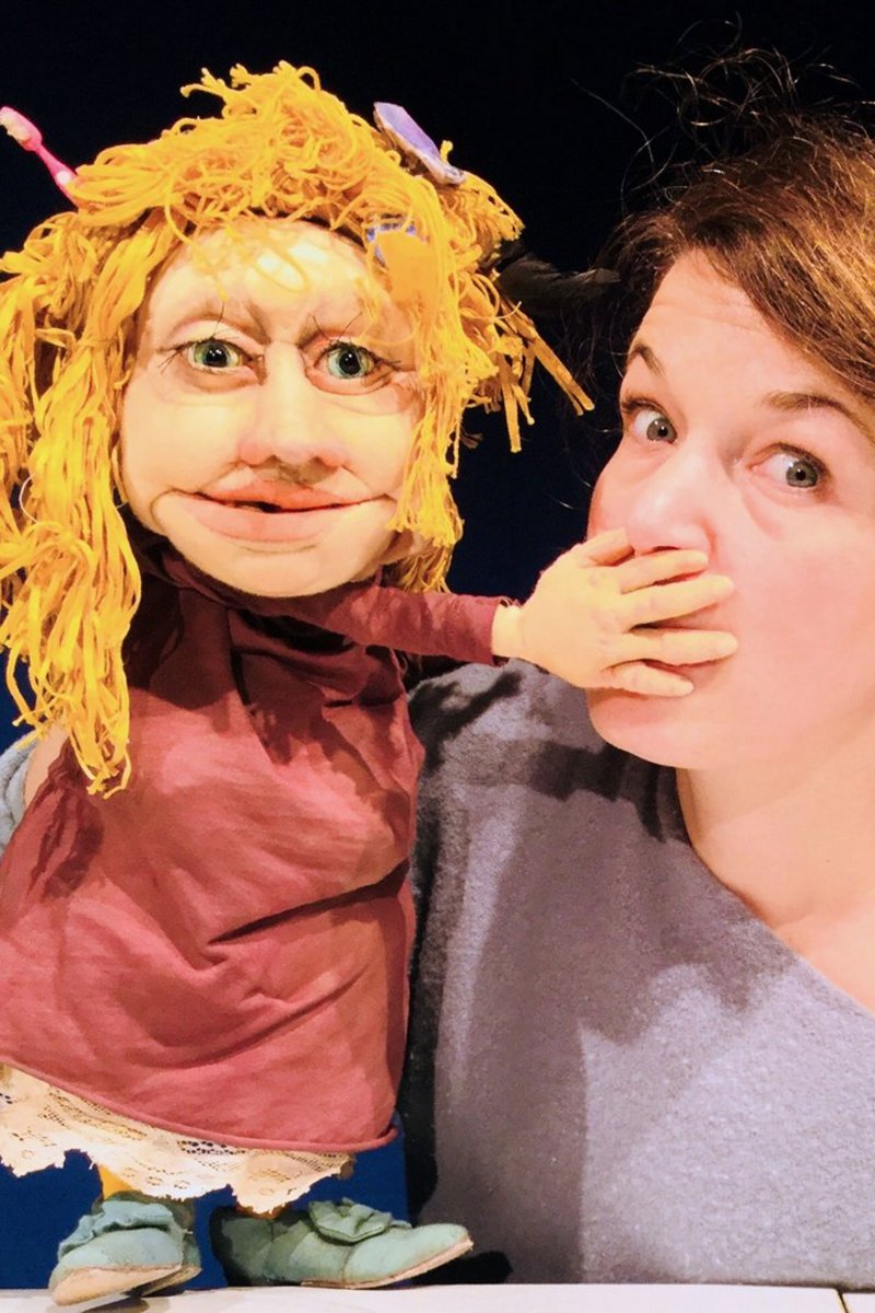 Rechts ist eine Frau mit heller Haut und dunklen Haaren, sie trägt einen grauen Pullover. Eine blonde Puppe in roter Kleidung hält der Frau den Mund zu.
