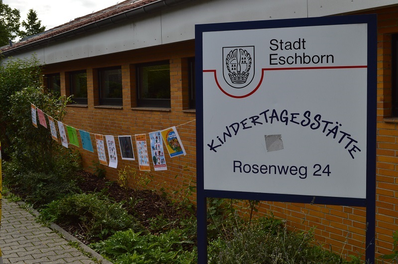 Neben dem weißen Schild mit der Aufschrift "Stadt Eschborn - Kindertagesstätte Rosenweg 24" hängen bunte Plakate an einer Schnur aufgereiht.