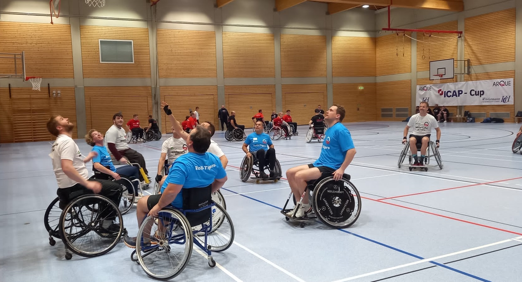 Rollstuhl-Basketball-Spielerinnen und -Spieler auf dem Feld. Sie tragen blaue, weiße und rote Trikots und schauen nach oben in Richtung Ball.