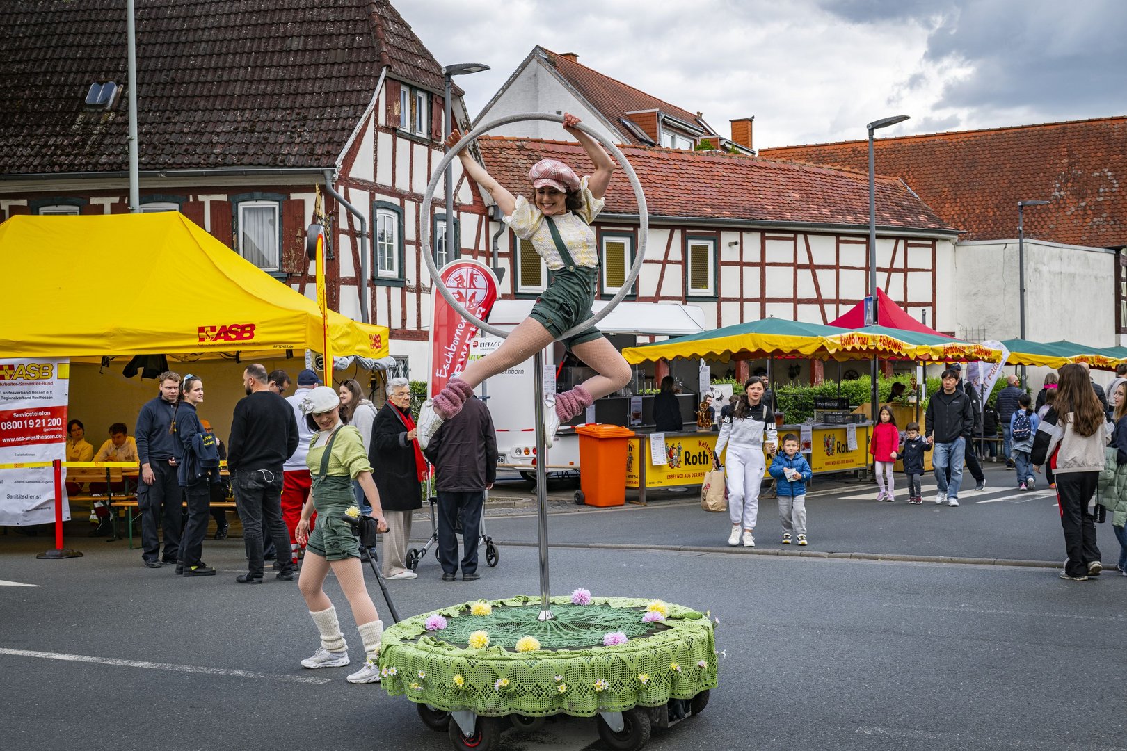 Eine Künstlerin von Flying Street Art zieht ein grünes Podest. Darauf ist ein Ring, in dem die andere Künstlerin sitzt. Beide tragen helle Ballonmützen und kurze grüne Latzhosen.