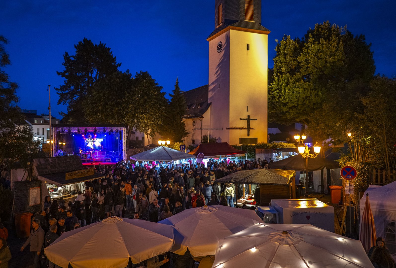 Das Eschenfest bei Nacht. Vor einem tiefblauen Himmel sieht man die weiße Fassade der veangelischen Kirche. Vorne sieht man weiße Schirme, dazwischen die vielen Menschen auf dem Eschenplatz.