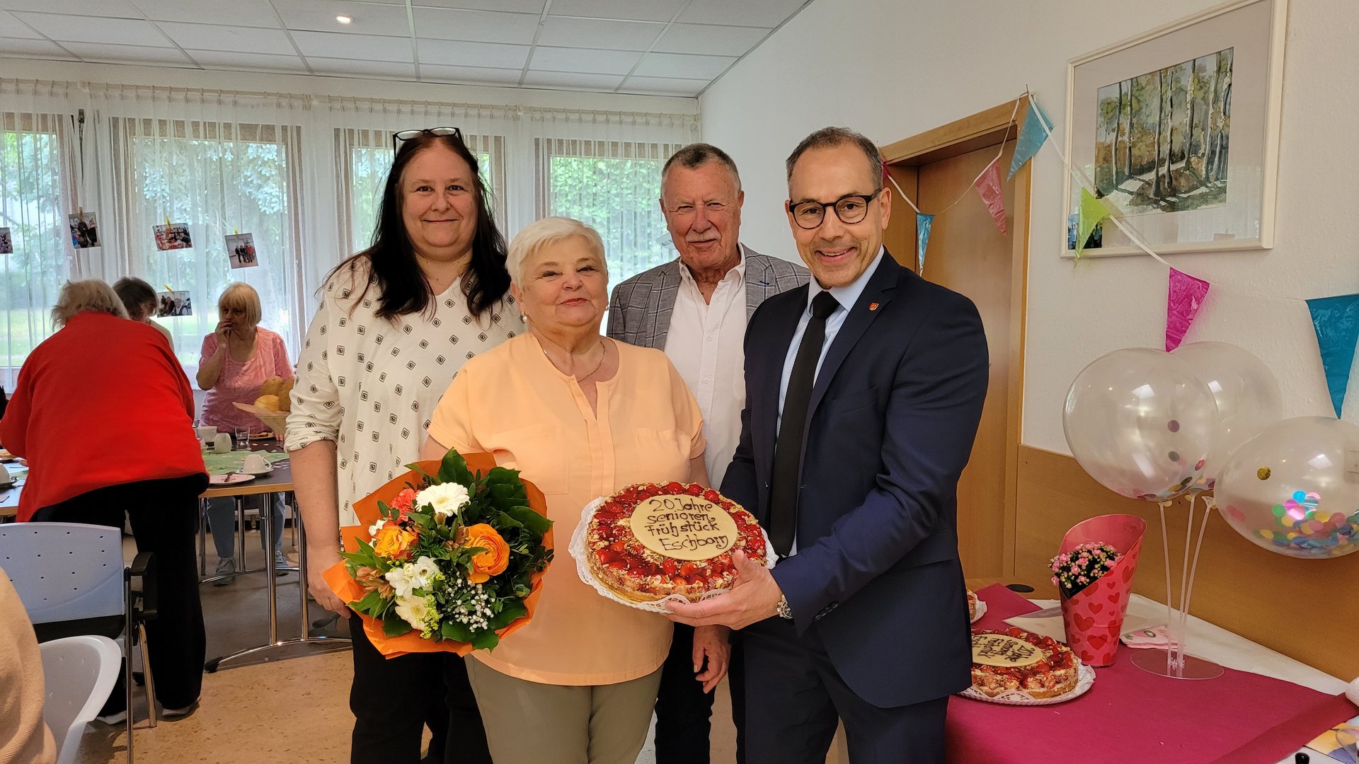 Katja Keiner, Anna Sabov mit einem Blumenstrauß, Helmut Bauch und Adnan Shaikh, der eine Erdbeertorte mit der Aufschrift "20 Jahre Seniorenfrühstück Eschborn" hält.