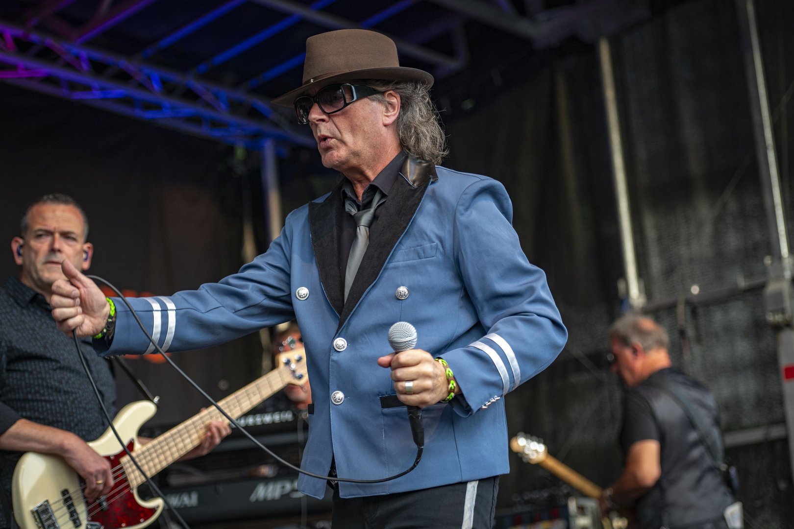 Karsten Bald als Udo-Lindenberg-Double. Er trägt den typischen schwarzen Hut, eine Sonnenbrille und ein blaues Sakko. In der Hand hält er ein Mikrofon.